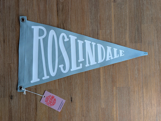 A light blue triangular cotton canvas pennant flag with 'ROSLINDALE' in large white vinyl lettering, laying on a brown wooden table. A small pink tag is attached near the left corner.