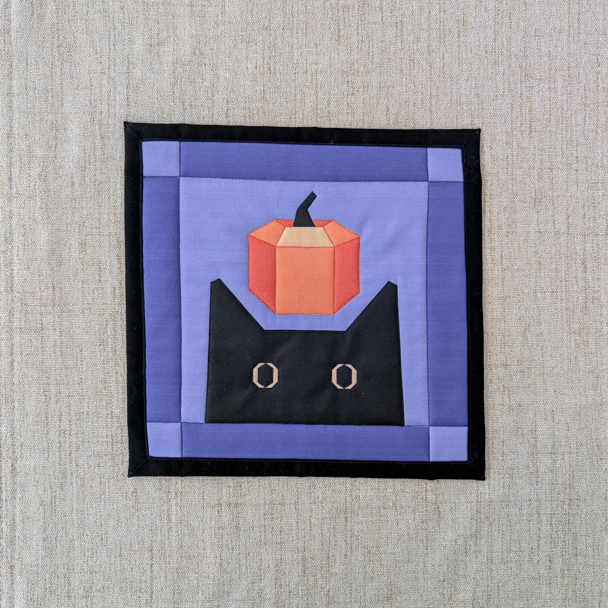 A square fabric wall hanging or quilted patch featuring a stylized black cat with a small orange pumpkin on its head, set against a purple background with a black border, displayed on a textured beige linen surface.