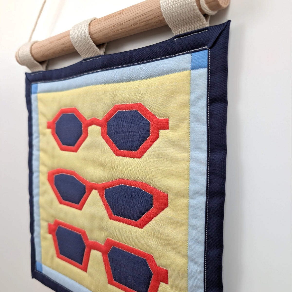 A small quilted banner with an illustration of three pairs of red sunglasses on a yellow background with a blue border, and is secured to 3 canvas straps around a wooden dowel, which is then hangs from a cream colored cotton string.