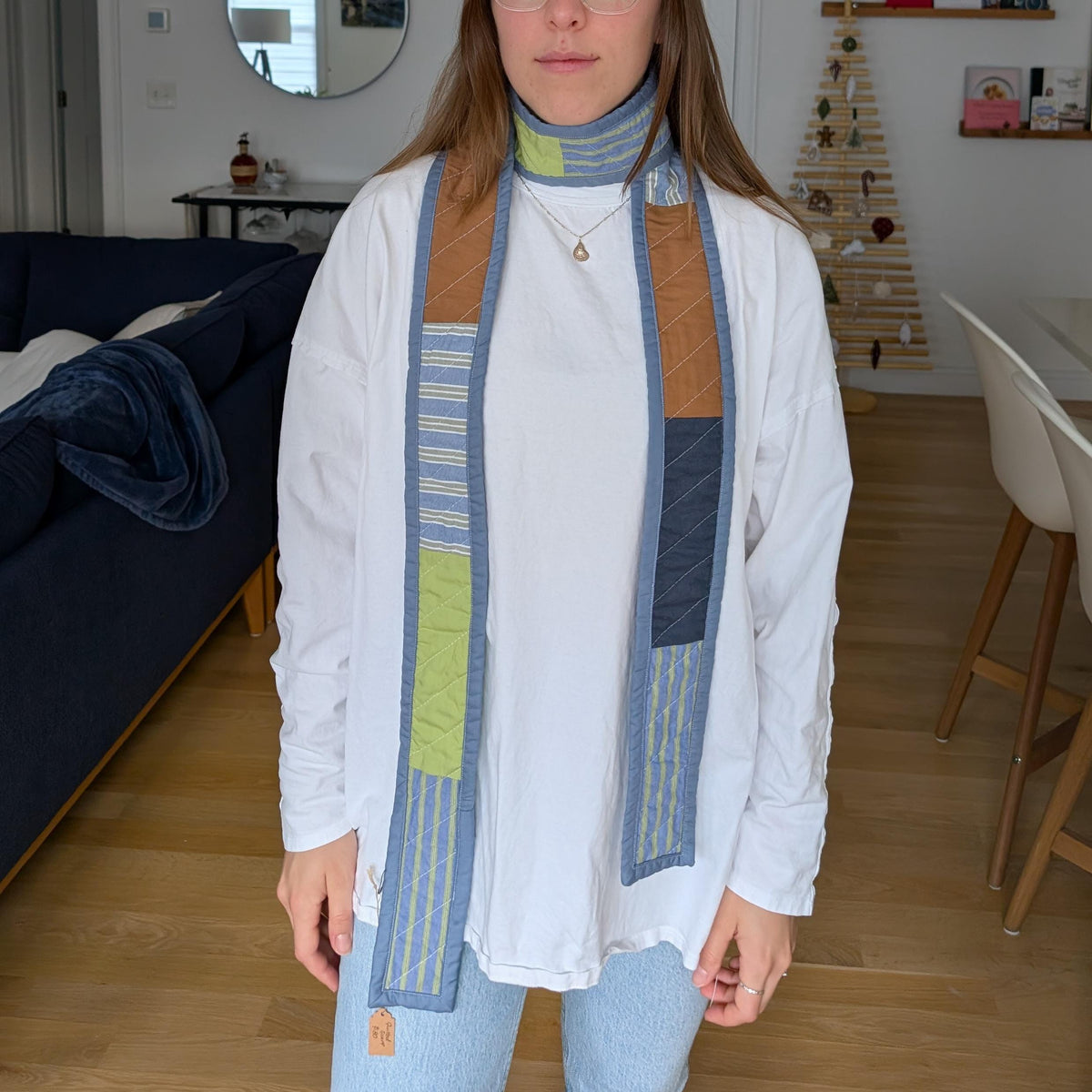 A woman wearing a white long-sleeve tunic top with a colorful quilted scarf draped around her neck, standing in a modern living room with wooden floors.