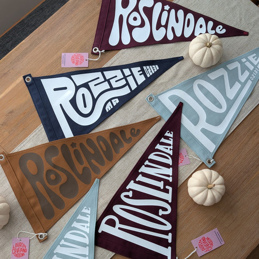 Six triangular fabric pennant flags with stylized 'ROZZIE' and 'ROSLINDALE' lettering in different colors and patterns, arranged on a wooden dining table with small white pumpkins and pink tags.