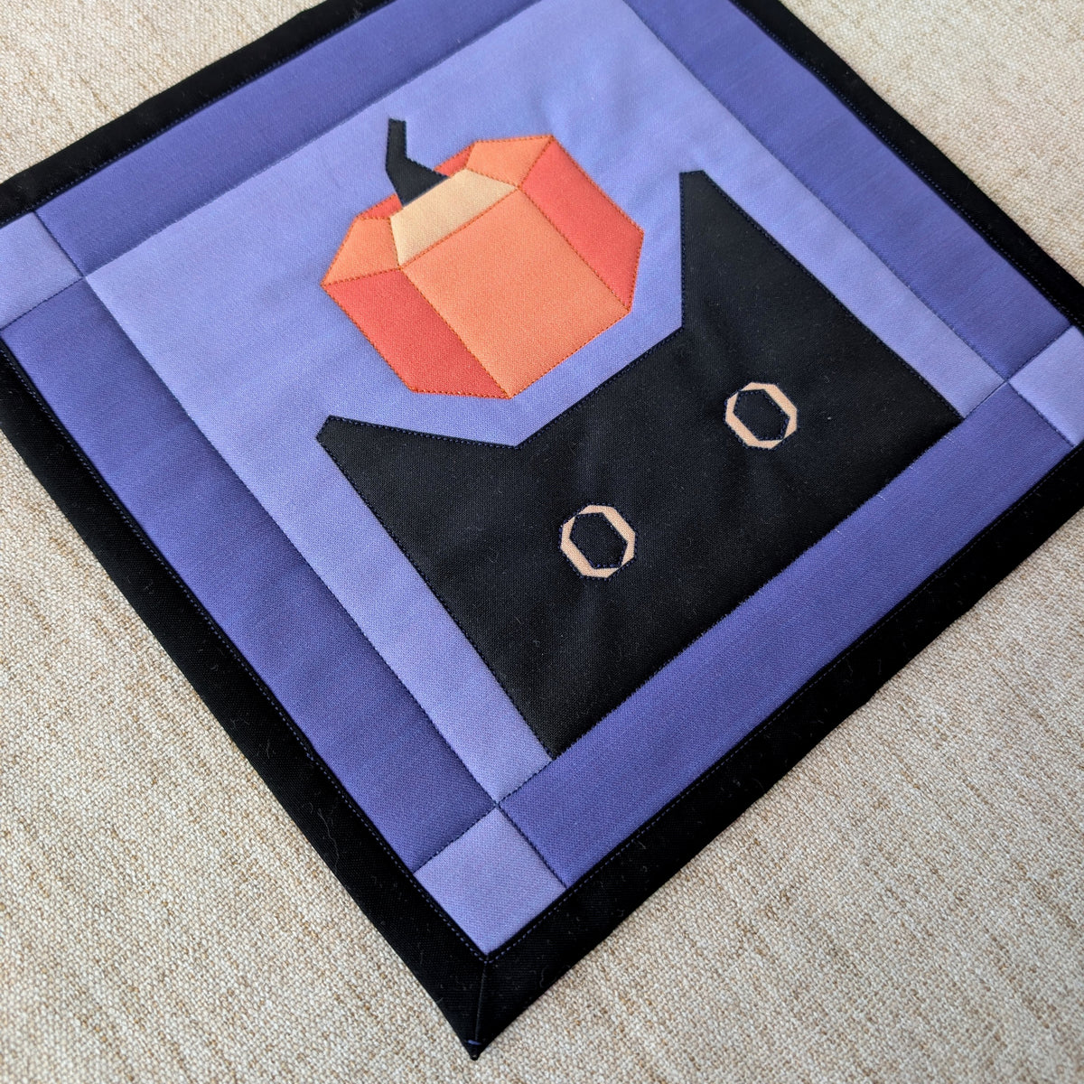 A square quilted fabric art piece featuring a stylized black cat with white eyes and a geometric orange pumpkin on a lavender and purple background, laid flat on a beige textured surface.