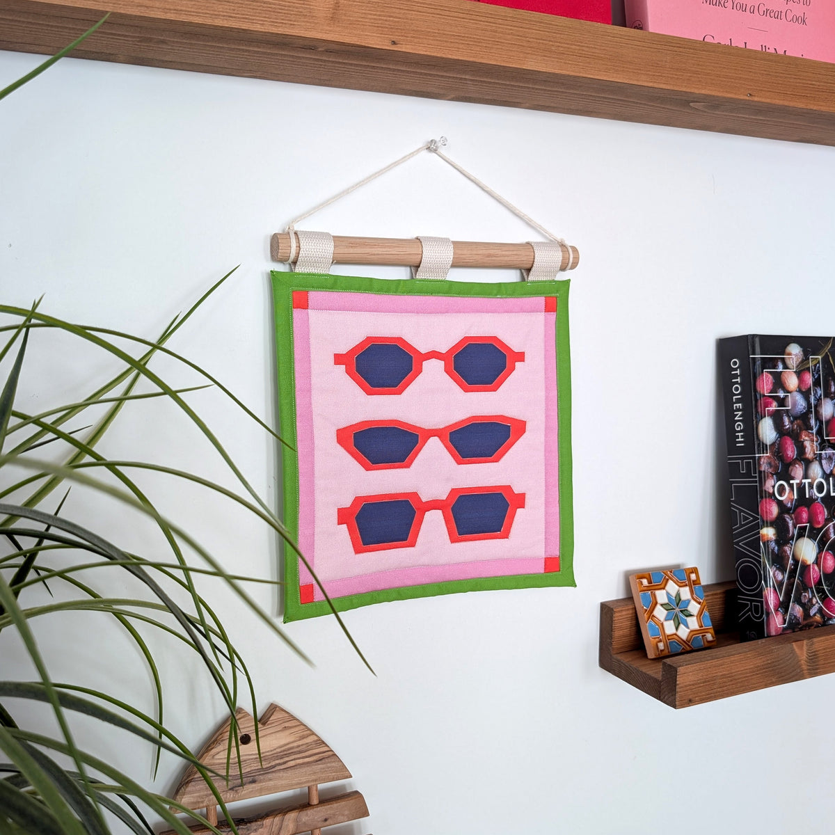 A small quilted banner with an illustration of three pairs of red sunglasses on a pink background with a green border, and is secured to 3 canvas straps around a wooden dowel, which is then hangs from a cream colored cotton string. Hanging on a colorful and busy kitchen wall.