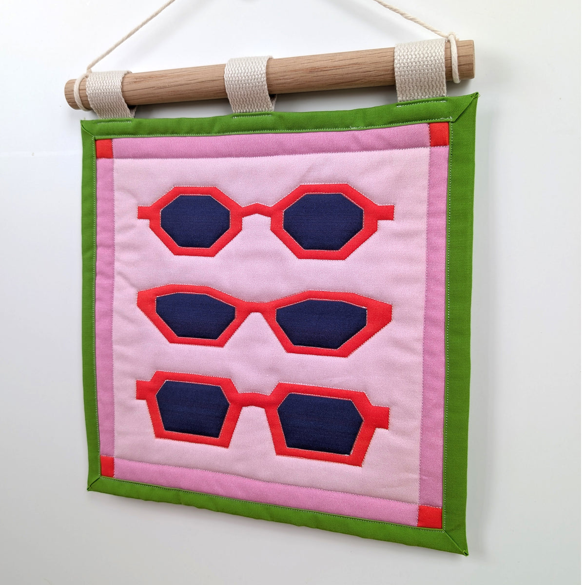 A small quilted banner with an illustration of three pairs of red sunglasses on a pink background with a green border, and is secured to 3 canvas straps around a wooden dowel, which is then hangs from a cream colored cotton string.