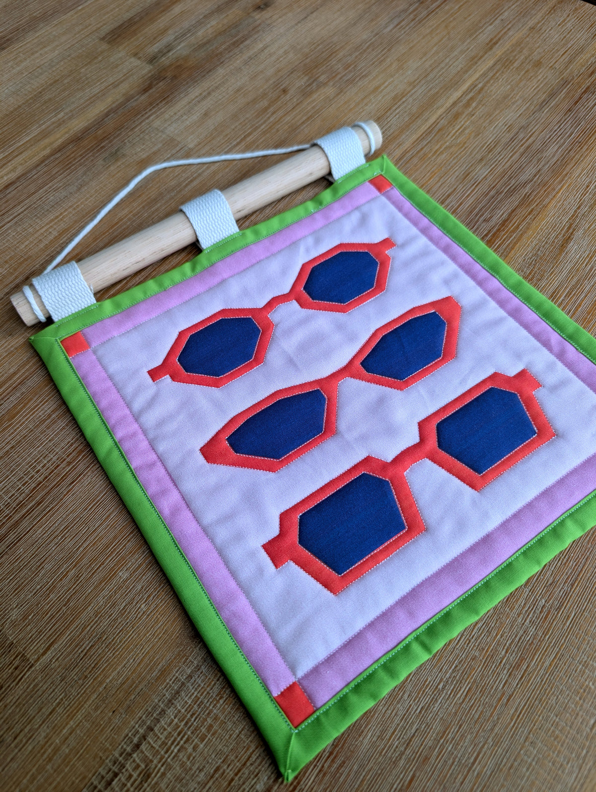 A small quilted banner with an illustration of three pairs of red sunglasses on a pink background with a green border, and is secured to 3 canvas straps around a wooden dowel, which is then hangs from a cream colored cotton string.