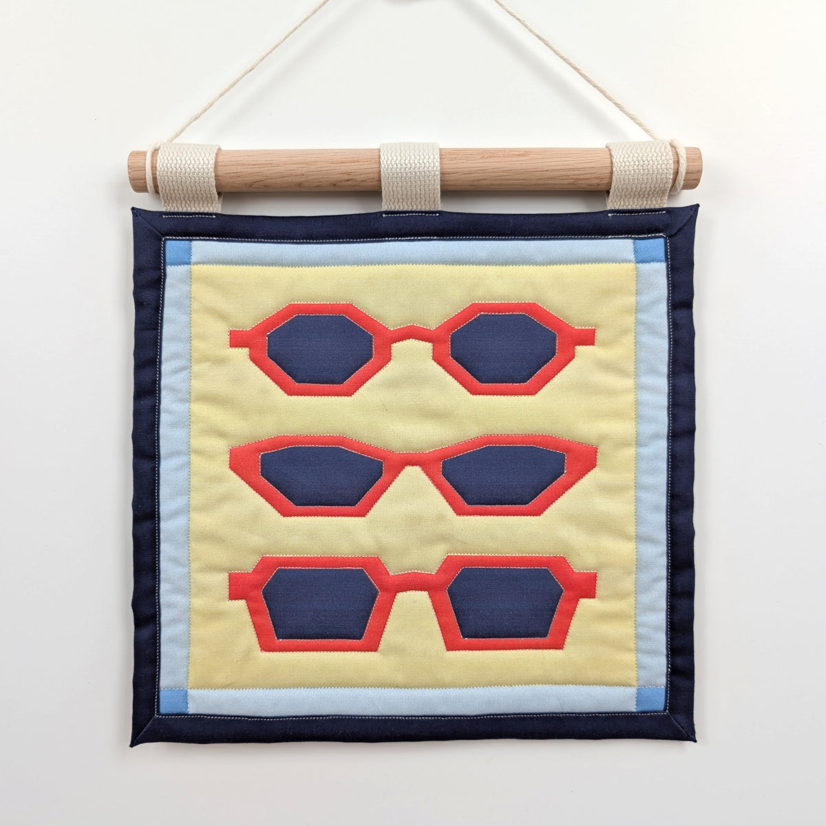 A small quilted banner with an illustration of three pairs of red sunglasses on a yellow background with a blue border, and is secured to 3 canvas straps around a wooden dowel, which is then hangs from a cream colored cotton string.