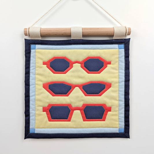 A small quilted banner with an illustration of three pairs of red sunglasses on a yellow background with a blue border, and is secured to 3 canvas straps around a wooden dowel, which is then hangs from a cream colored cotton string.