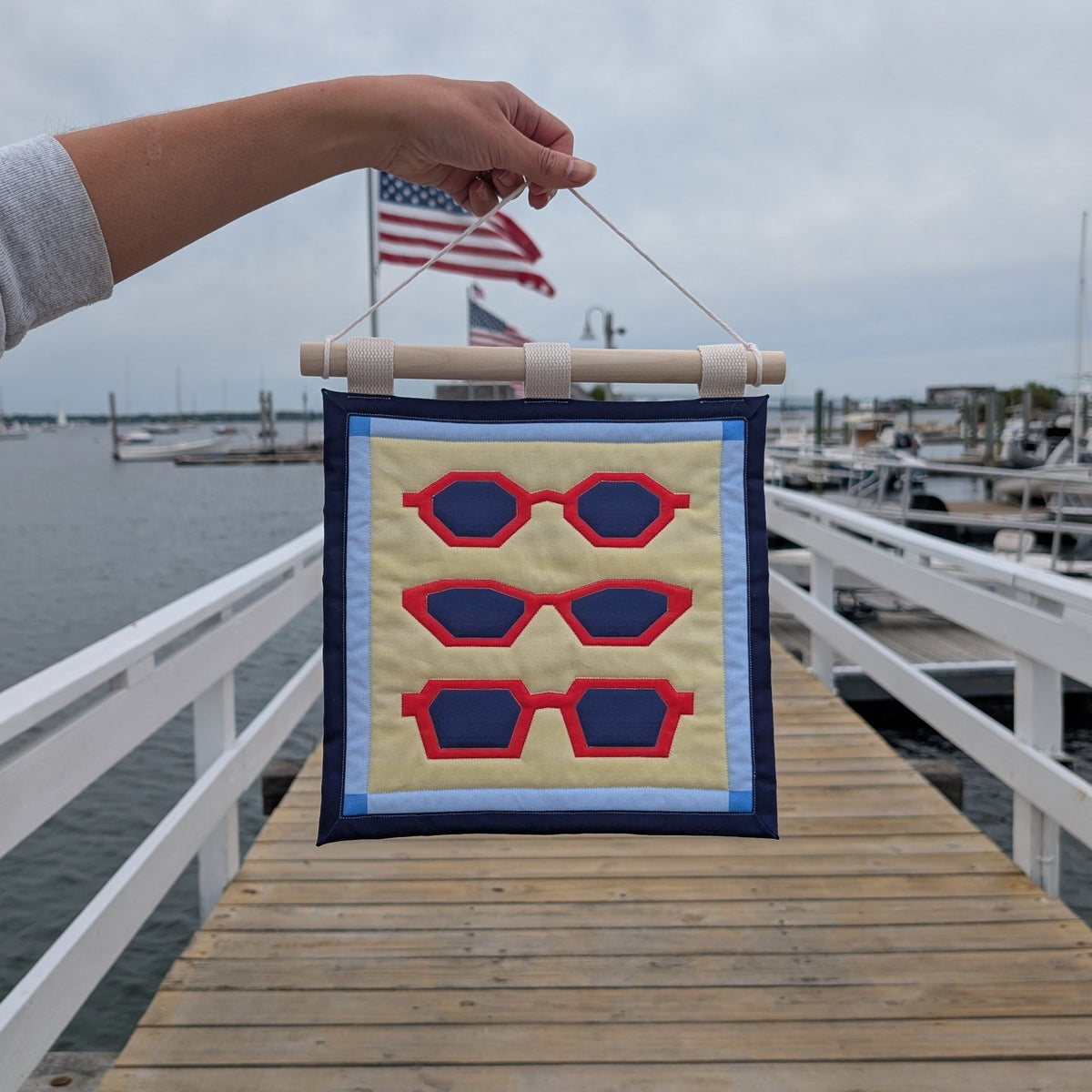 An outstretched arm holds a small quilted banner over a wooden dock on the ocean, with boats and an overcast sky in the background. The banner features three pairs of red sunglasses on a yellow background with a blue border, and is secured to 3 canvas straps around a wooden dowel, which is then hangs from a cream colored cotton string.