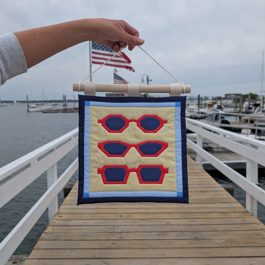 An outstretched arm holds a small quilted banner over a wooden dock on the ocean, with boats and an overcast sky in the background. The banner features three pairs of red sunglasses on a yellow background with a blue border, and is secured to 3 canvas straps around a wooden dowel, which is then hangs from a cream colored cotton string.