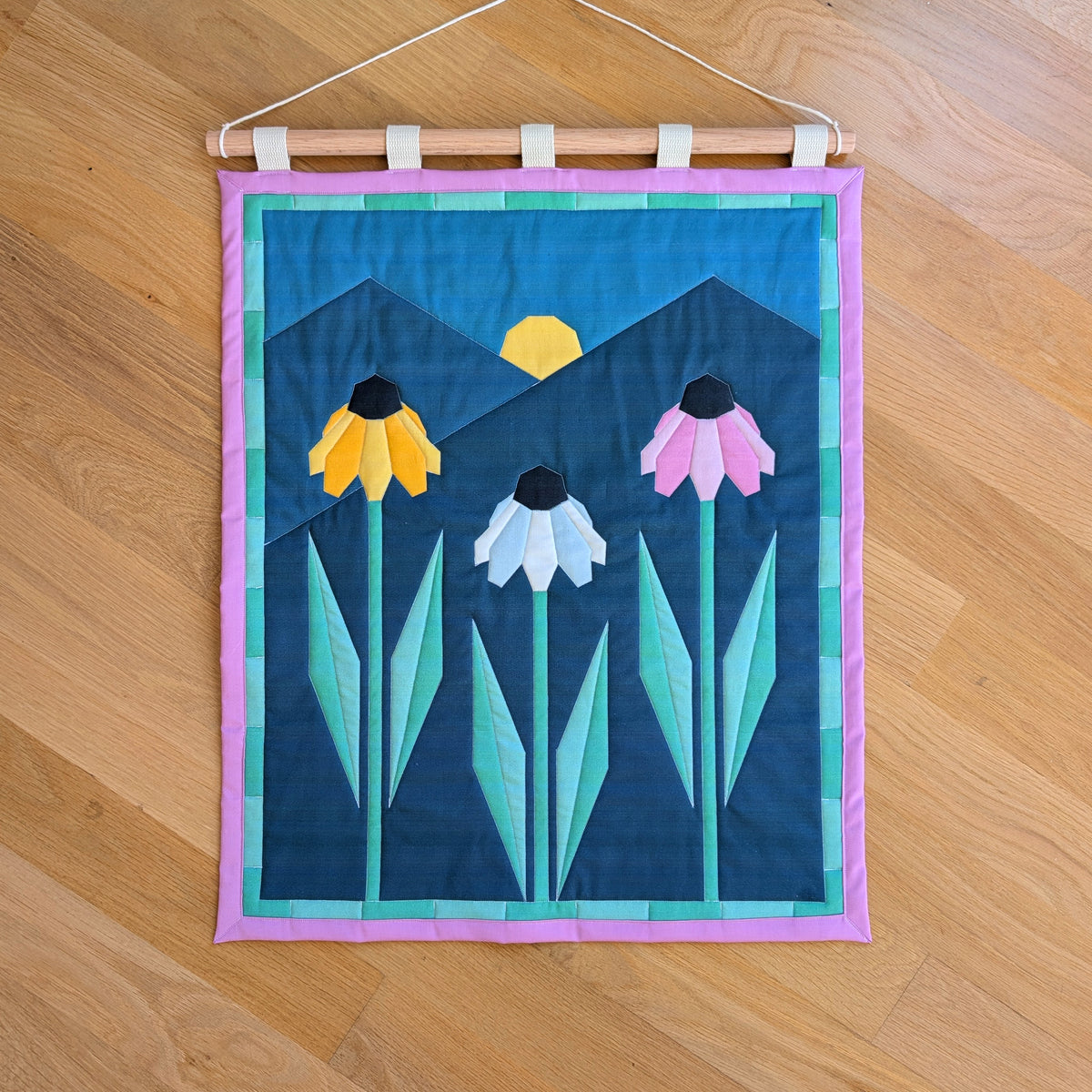 A quilted banner with an illustration of three echinacea flowers in front of 2 mountains with the sun rising behind them. It is secured to 5 canvas straps around a wooden dowel, which then hangs from a cream colored cotton string.