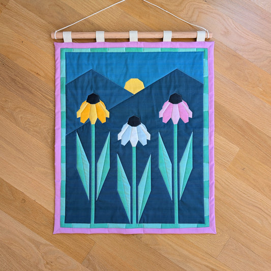 A quilted banner with an illustration of three echinacea flowers in front of 2 mountains with the sun rising behind them. It is secured to 5 canvas straps around a wooden dowel, which then hangs from a cream colored cotton string.