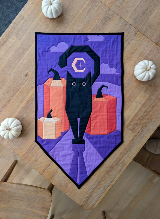 A quilted wall hanging with a pointed bottom and an angular illustration of a black cat standing in front of three orange pumpkins, with a crescent moon and clouds in a purple night sky behind them.